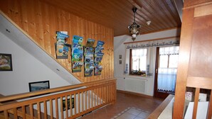Interior de madera con una escalera, un tablón de anuncios con folletos de viajes y una ventana con cortinas decorativas.