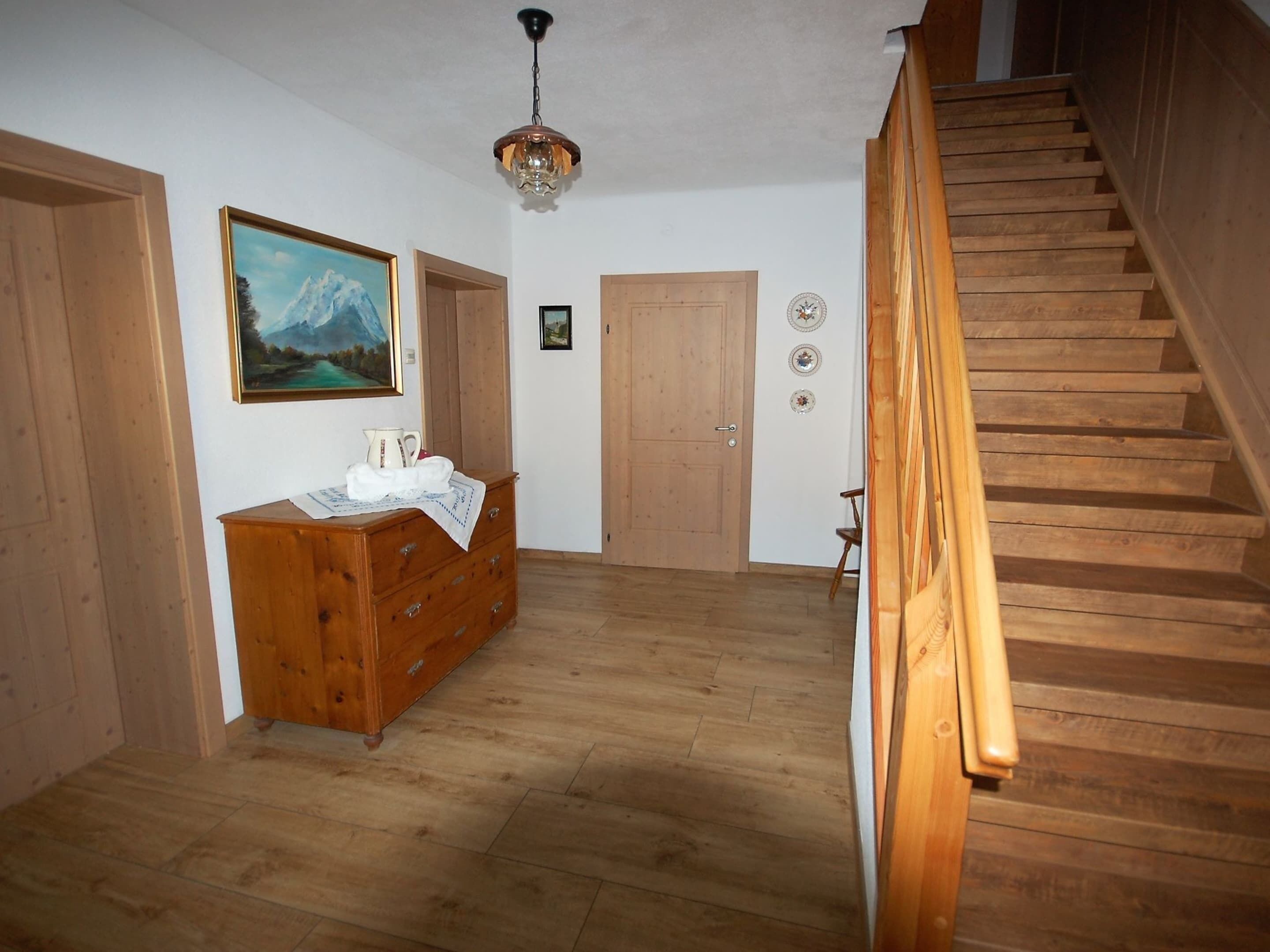 A wooden staircase leading to an upper floor, a wooden dresser with a painting, and a door with decorative plates.