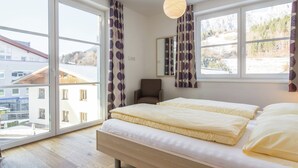 A bedroom with a large bed, two windows, and a view of buildings and mountains. - Apartment in Leogang Near Ski Slopes (Leogang)