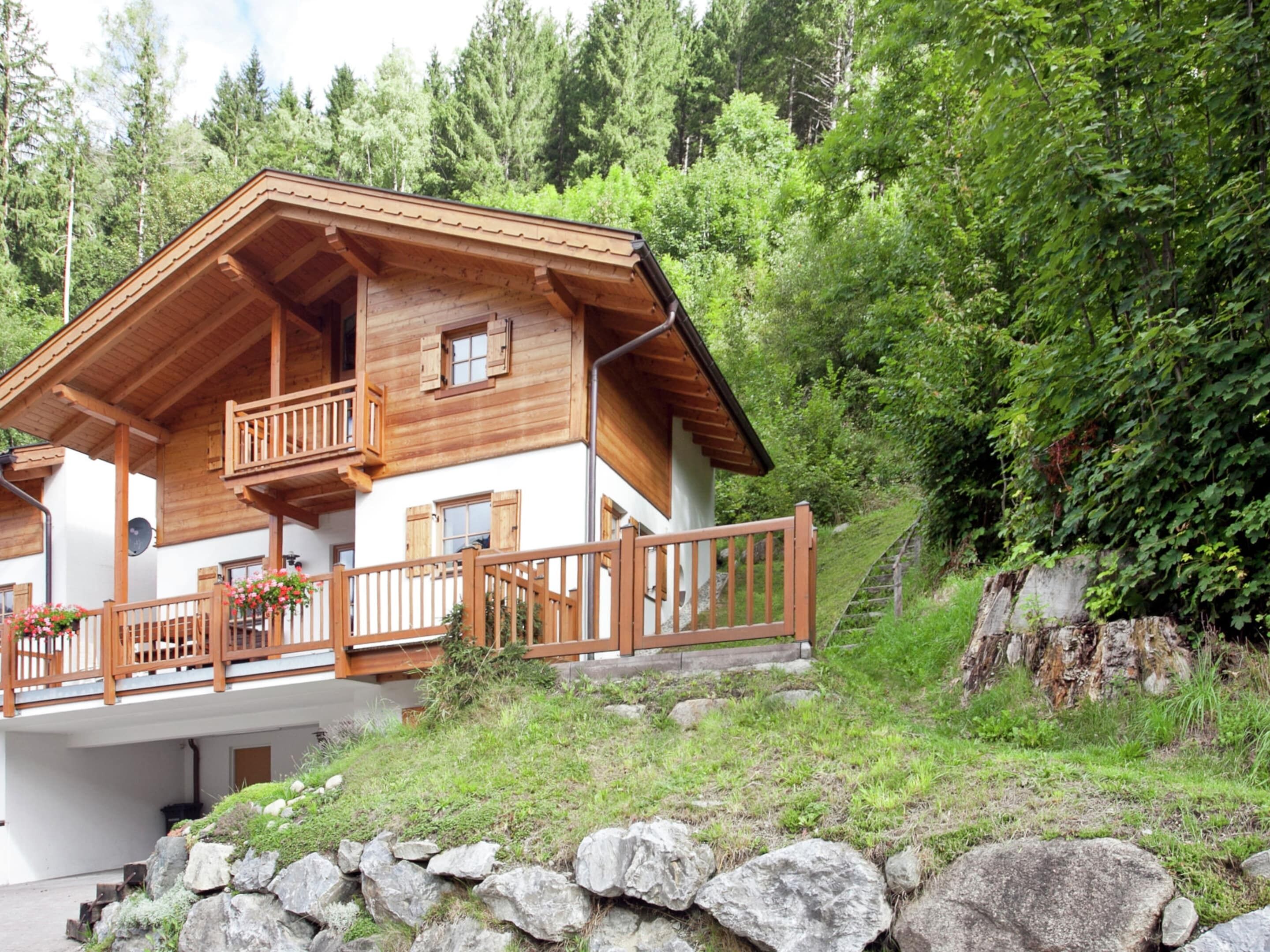 Een houten chalet met balkon, omgeven door weelderig groen en een rotsachtige heuvel.