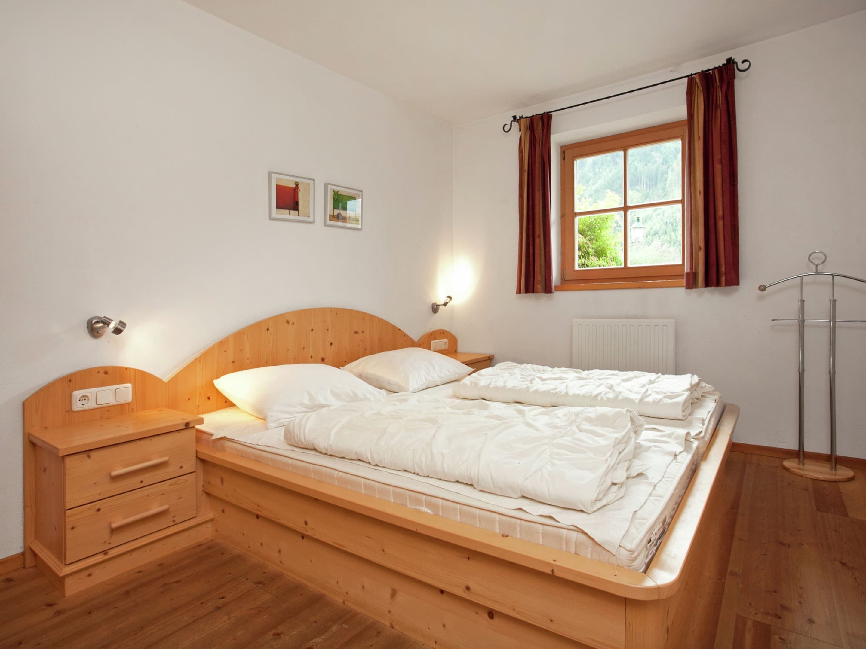 Un dormitorio con cama de madera, mesitas de noche, radiador, ventana con cortinas y dos cuadros en la pared.