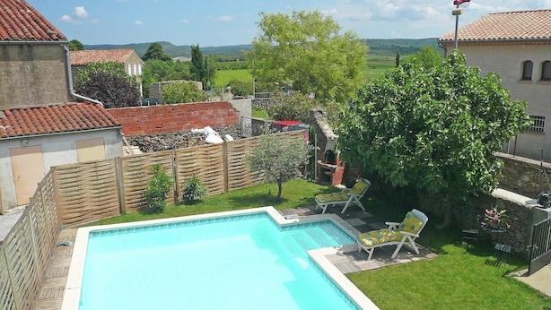 A swimming pool with a wooden deck and lounge chairs, surrounded by a fenced yard with a view of a rural landscape. - Holiday Home Talairan With Pool and BBQ (Talairan)
