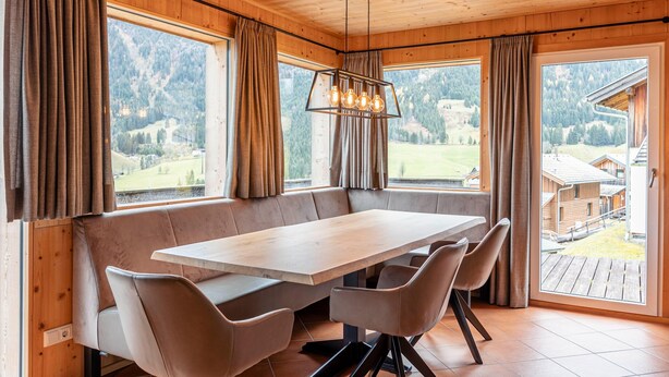 Interior de una cabaña de madera con una gran mesa de comedor, bancos acolchados y ventanas amplias que ofrecen vistas a la montaña.