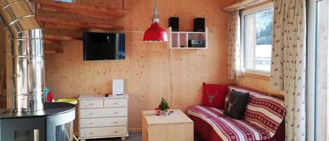 L’intérieur chaleureux d’un chalet avec un poêle à bois, un canapé agrémenté de coussins à motifs, un téléviseur et une étagère en bois où sont disposés des objets décoratifs.