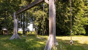Ein hölzernes Schaukelgestell in einer grünen Wiese mit Bäumen im Hintergrund.