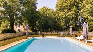 Ein rechteckiger Swimmingpool mit klarem, blauem Wasser, umgeben von einem Holenzaun, Bäumen und Liegestühlen mit Sonnenschirmen.