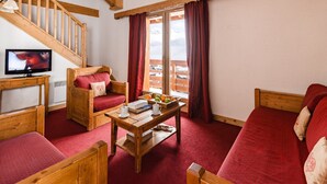 Ein gemütliches Zimmer mit einer Holztreppe, einem Fernseher, einem Sofa, einem Sessel und einem Tisch mit einem Buch und Obst.