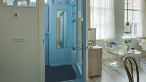 Une pièce avec une porte bleue, une chaise blanche avec un coussin bleu, une petite table avec un vase et un luminaire suspendu.