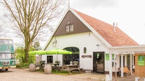 Un bâtiment blanc avec un toit brun, un autobus à deux étages vert garé devant, et des tables et chaises à l’extérieur, protégées par des parasols verts.