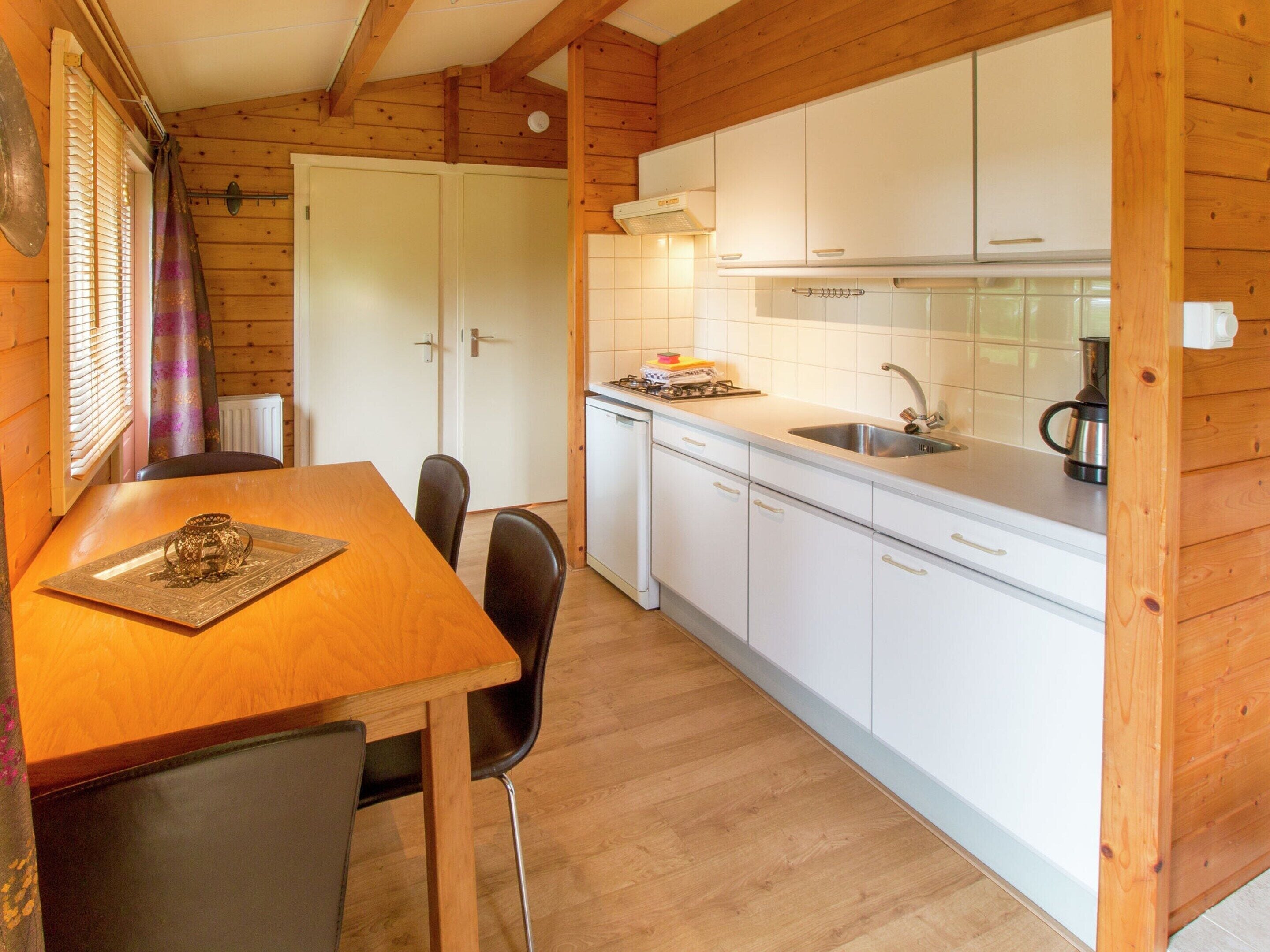 A wooden cabin interior with a dining table, chairs, a kitchen with white cabinets, and a window with blinds.