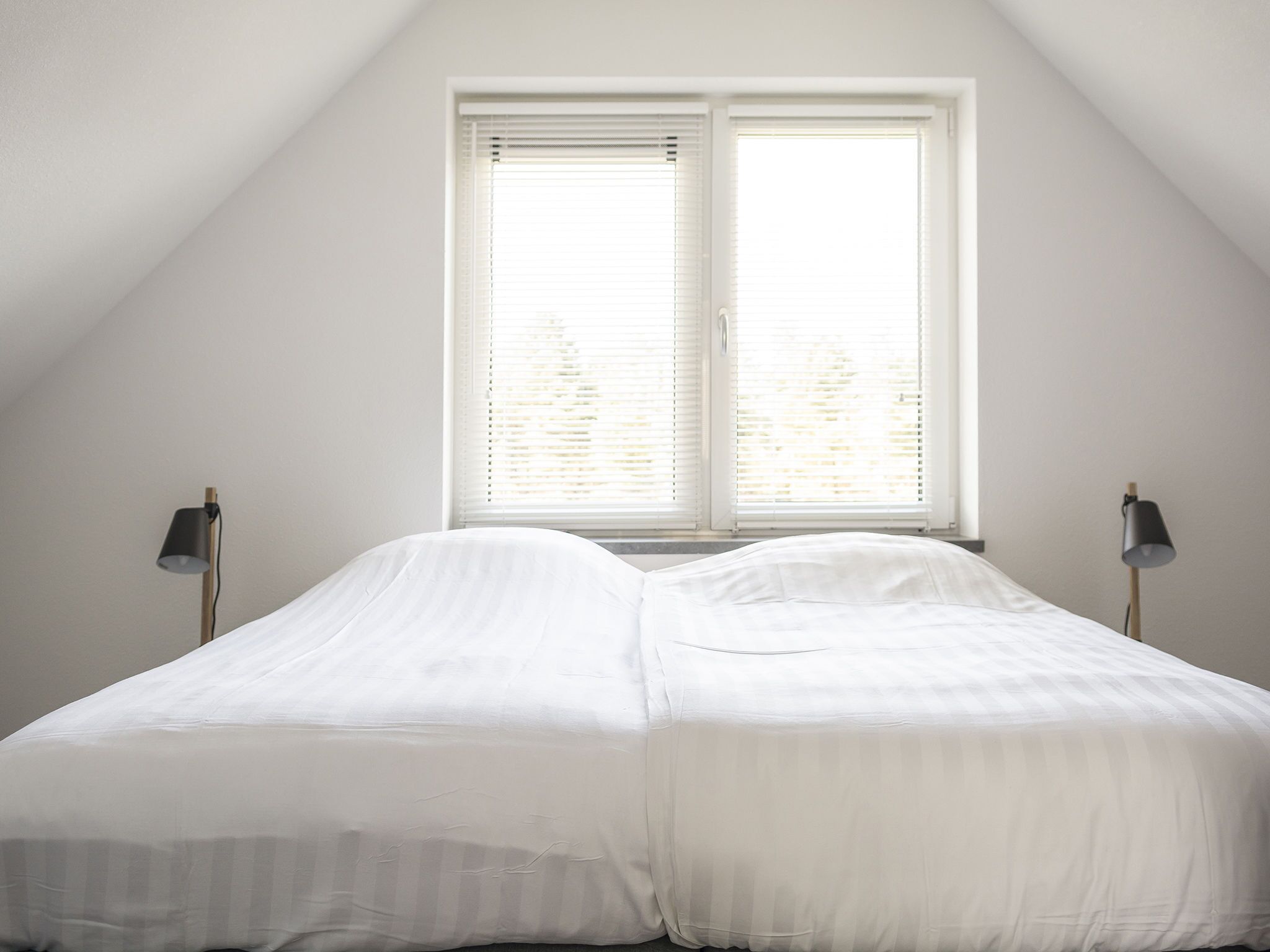A bedroom with a sloped ceiling, two bedside lamps, and a window with blinds.