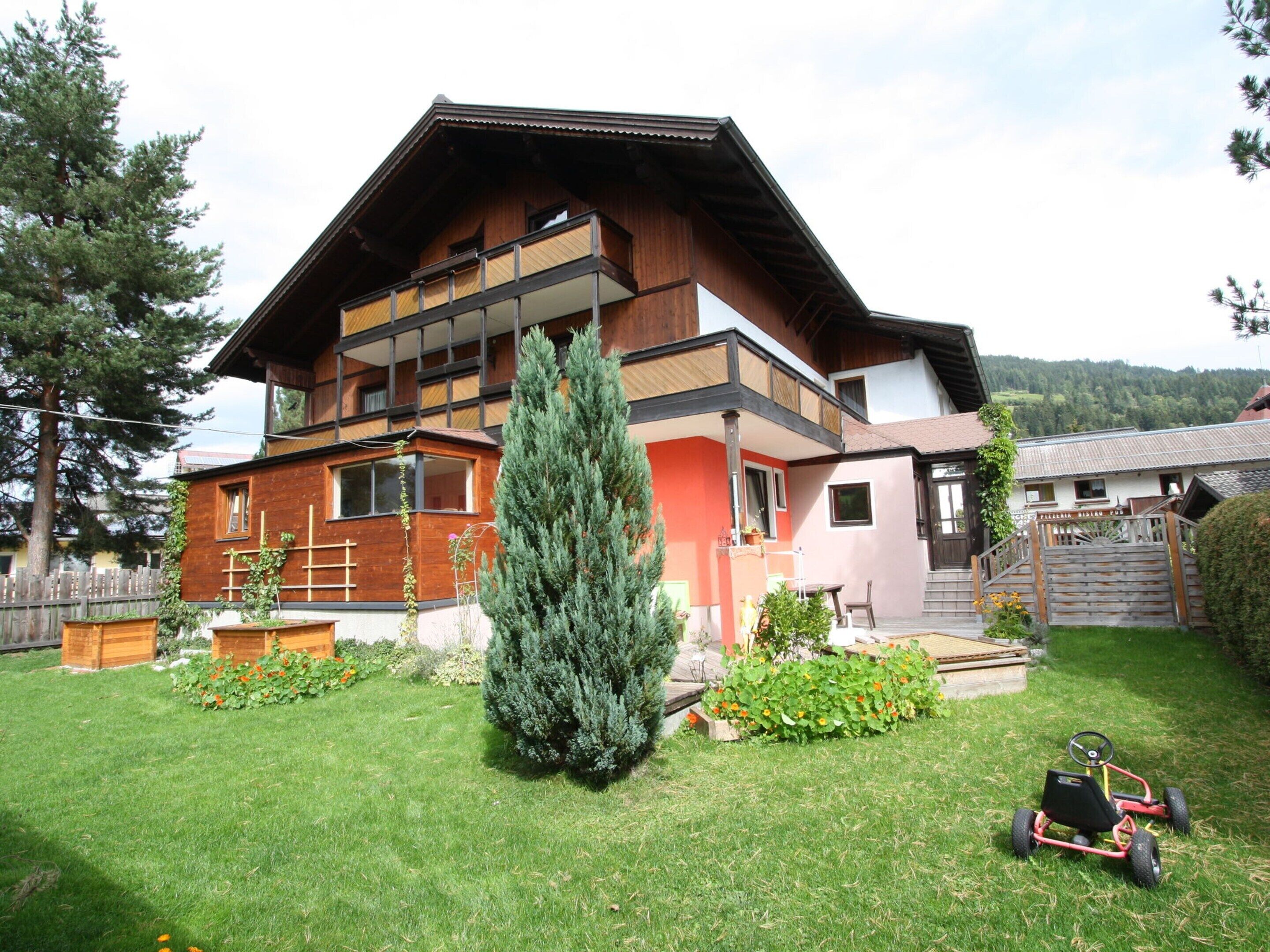 A two-story wooden house with a balcony, surrounded by a well-maintained lawn and a tall evergreen tree.