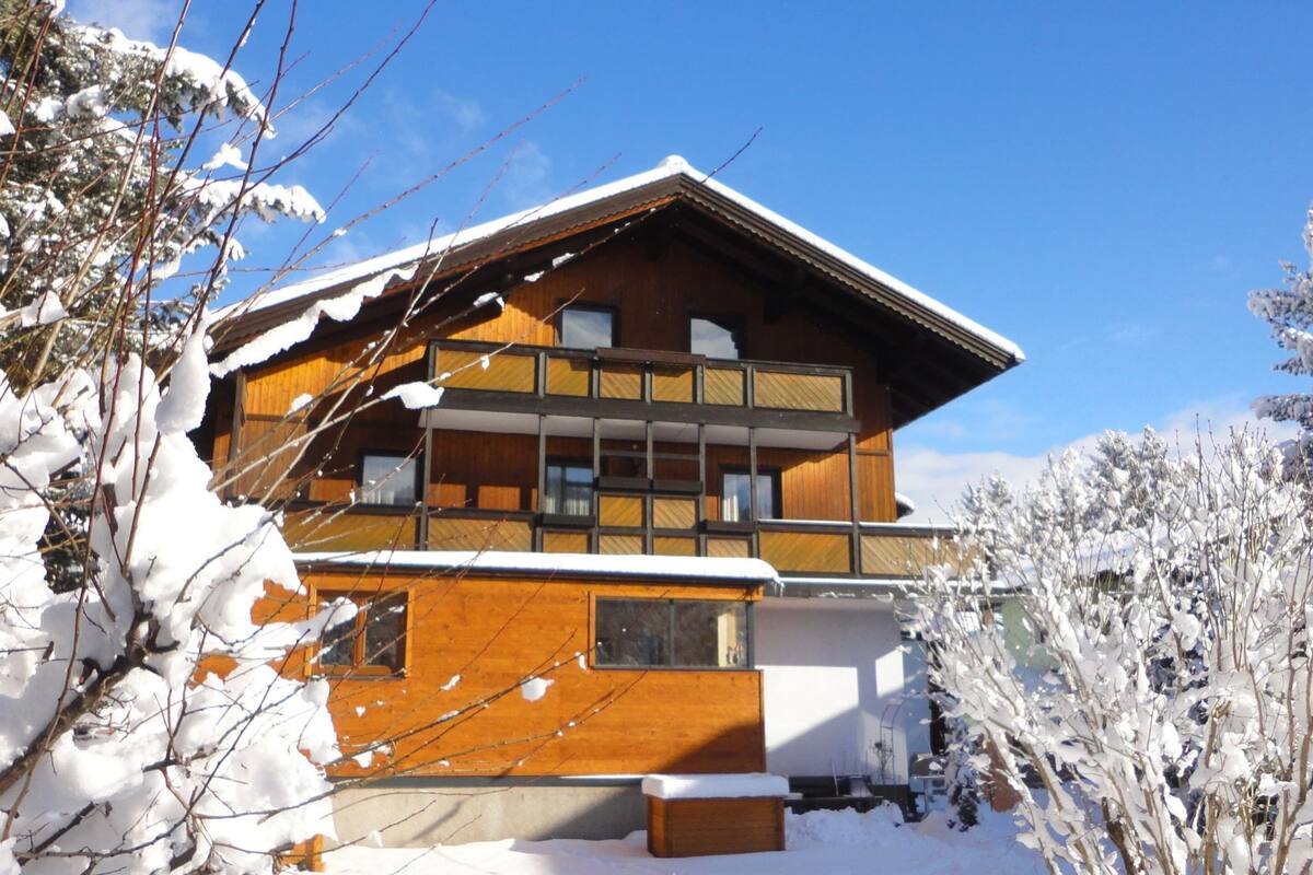 A two-story wooden chalet with a balcony, surrounded by snow-covered trees.