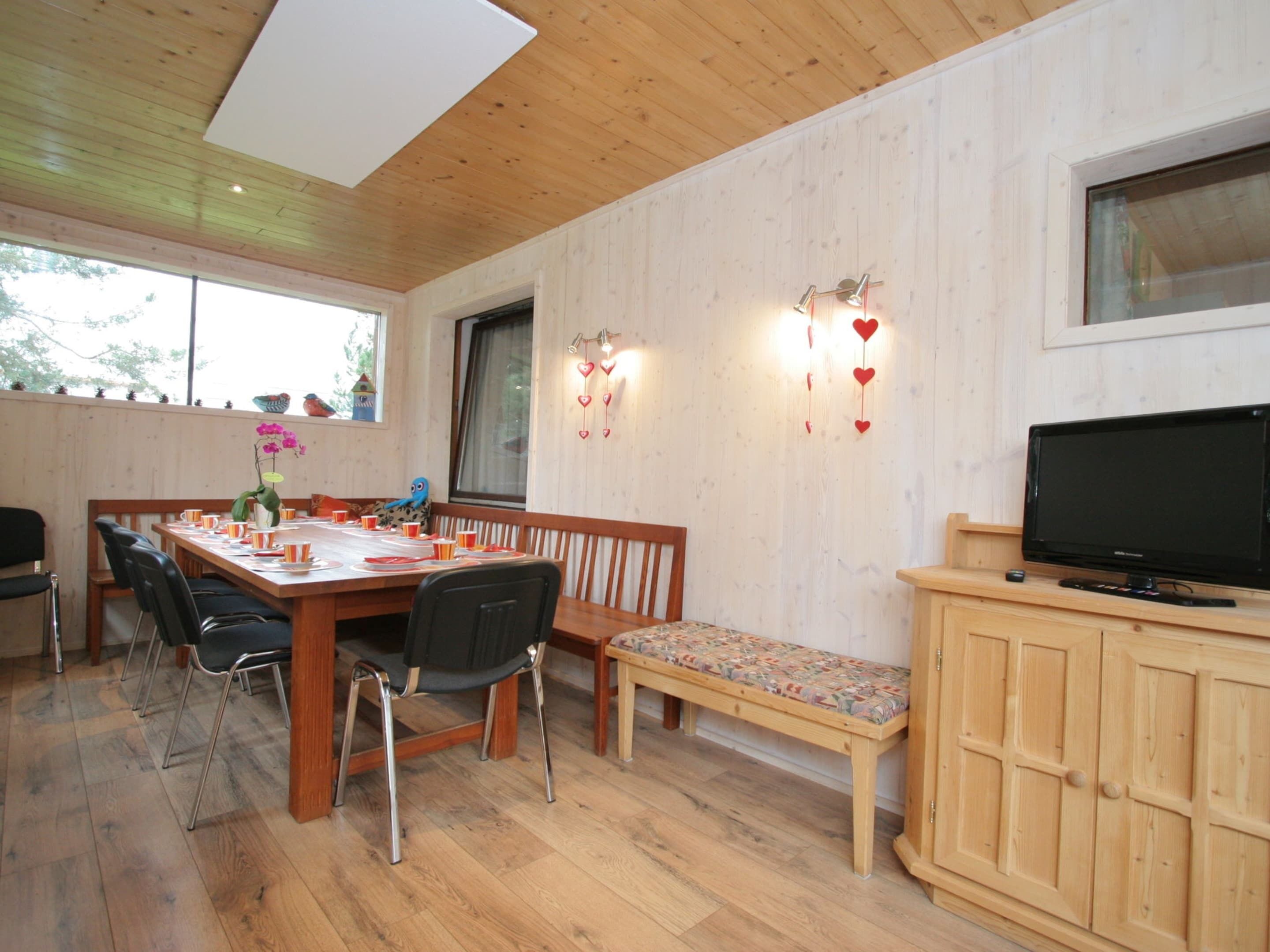 A dining area with a wooden table, chairs, and a bench. There is a television on a wooden cabinet.