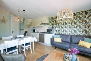 A modern kitchen with white cabinets, a dining area with a white table and chairs, a dark grey sofa with yellow cushions, and a floral wallpaper.