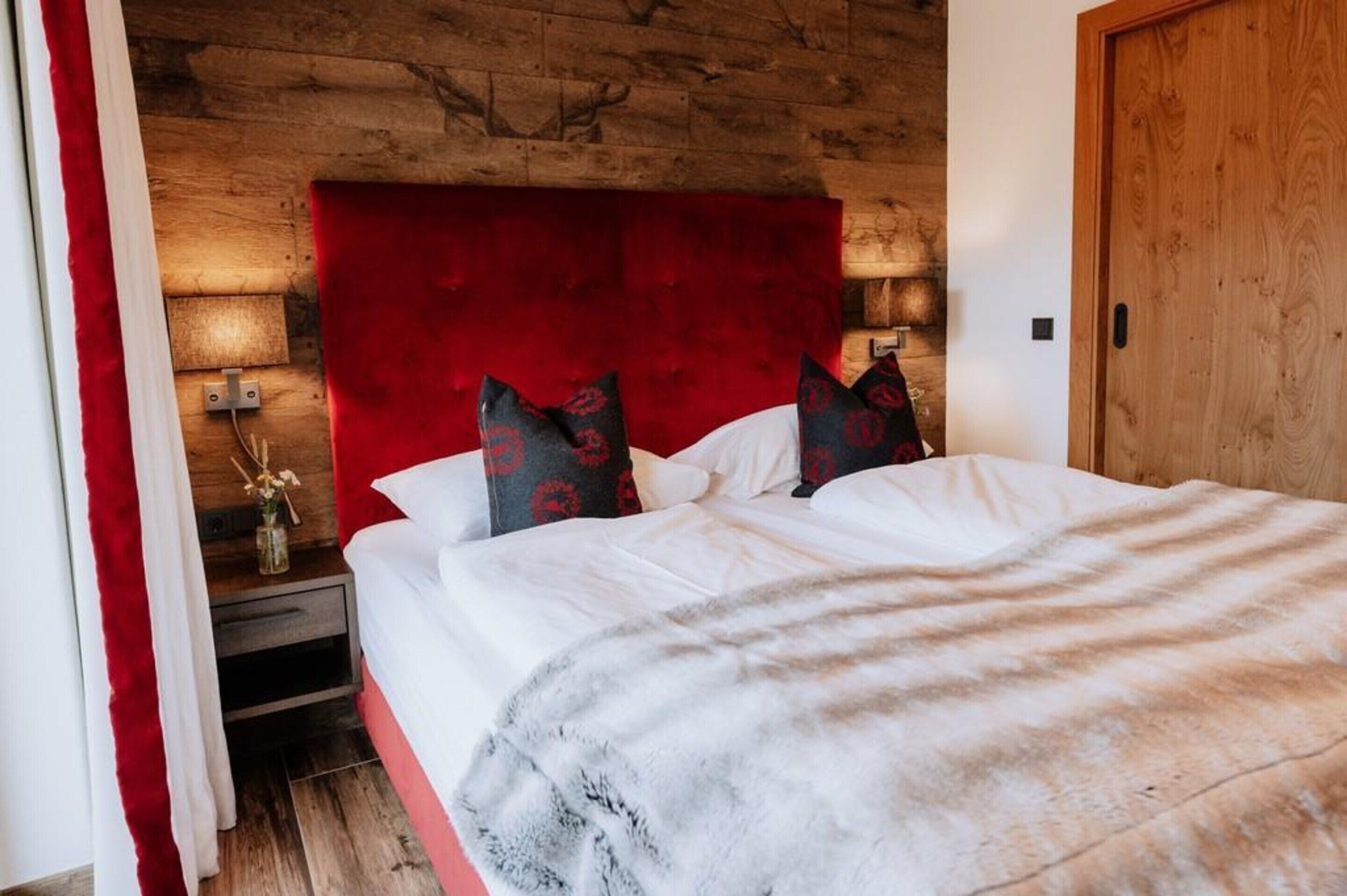 A hotel room with a wooden headboard, a red accent wall, and a bed with white linens and a grey blanket.