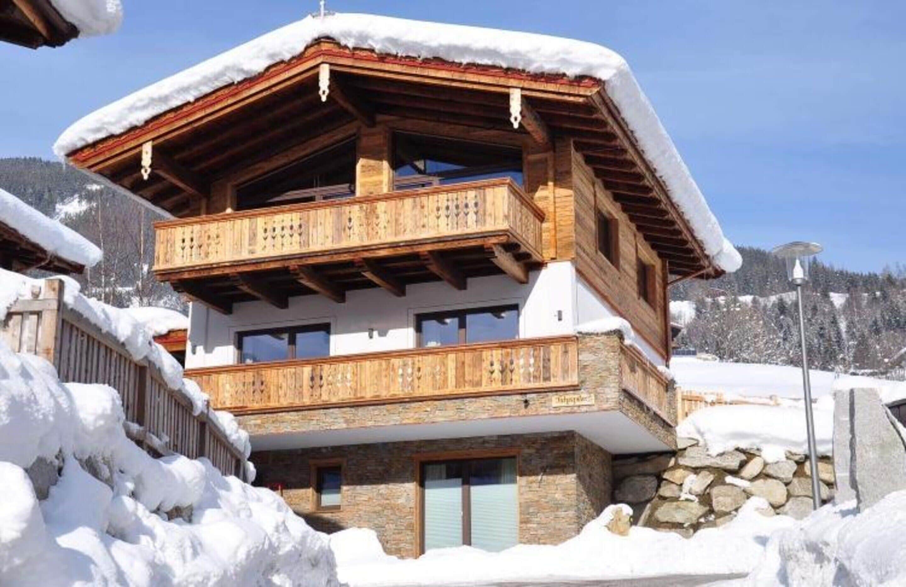 A two-story chalet with wooden balconies and a stone foundation, surrounded by snow.