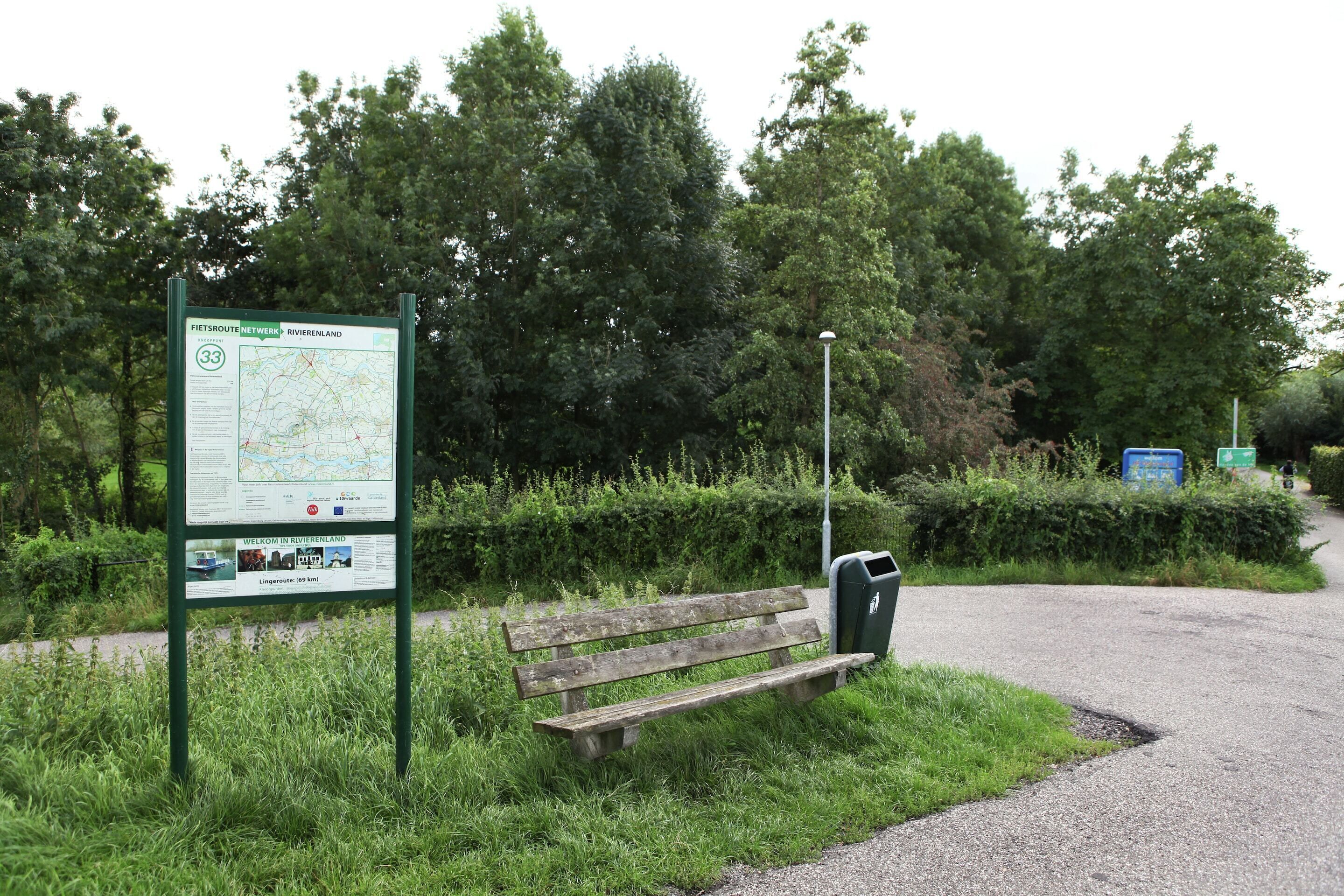 Eine Bank, ein Hinweisschild mit einer Karte und ein Laternenpfahl in einer Parkanlage.