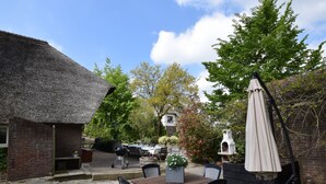 A patio with a table and chairs, a thatched-roof building, and a garden with trees and a birdhouse. - Quaint Farmhouse near River in Oosterwijk (Oosterwijk)