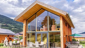 A modern wooden cabin with large glass windows, outdoor seating, and a view of the surrounding landscape.