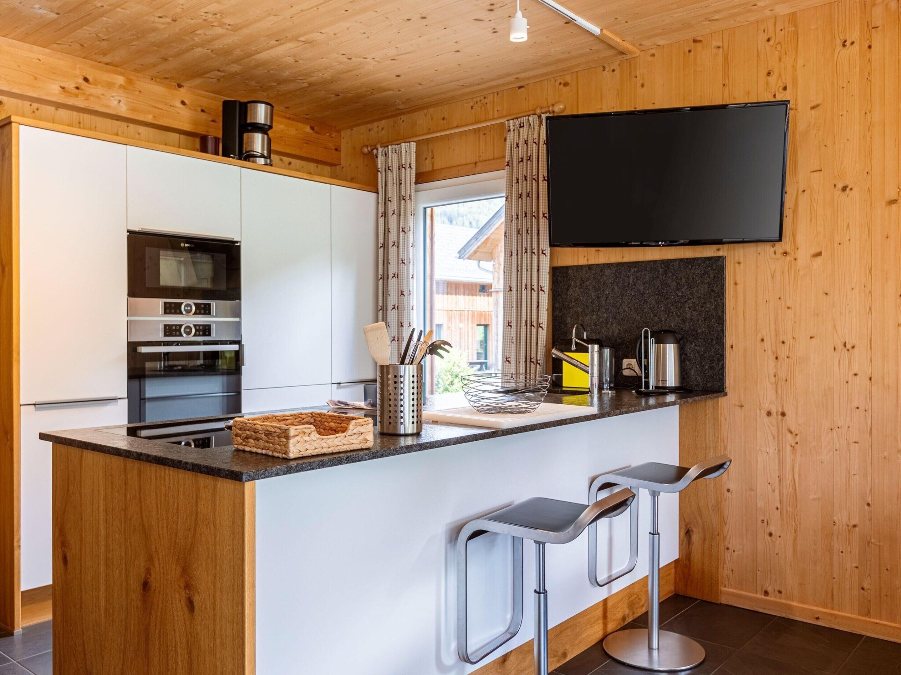 A modern kitchen with white cabinets, a built-in oven, a coffee machine, and a flat-screen TV mounted on the wall.