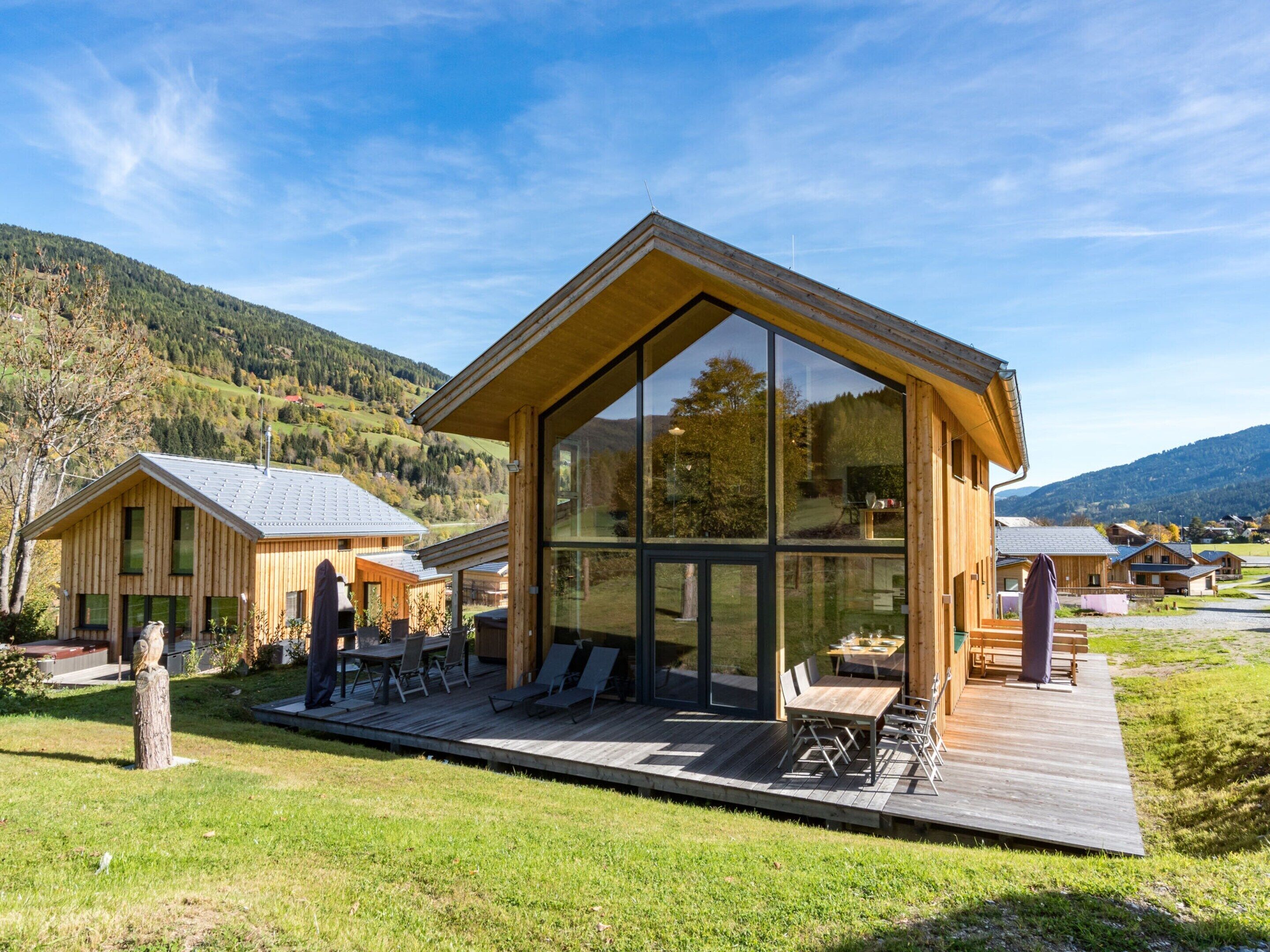 A modern wooden cabin with large glass windows, a deck, and a view of the surrounding hills.
