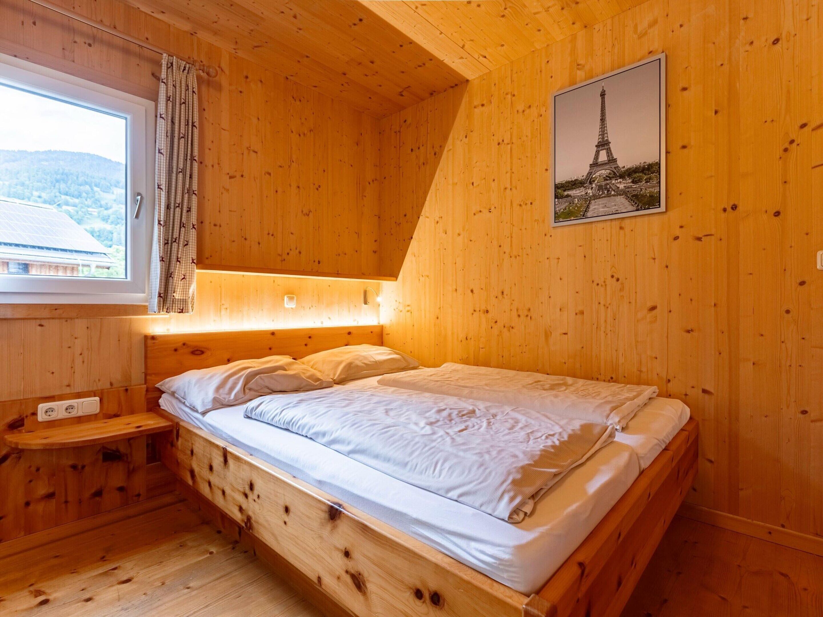 Une chambre rustique en bois, avec un lit, une fenêtre avec des rideaux, une table de chevet et un tableau représentant la Tour Eiffel accroché au mur.