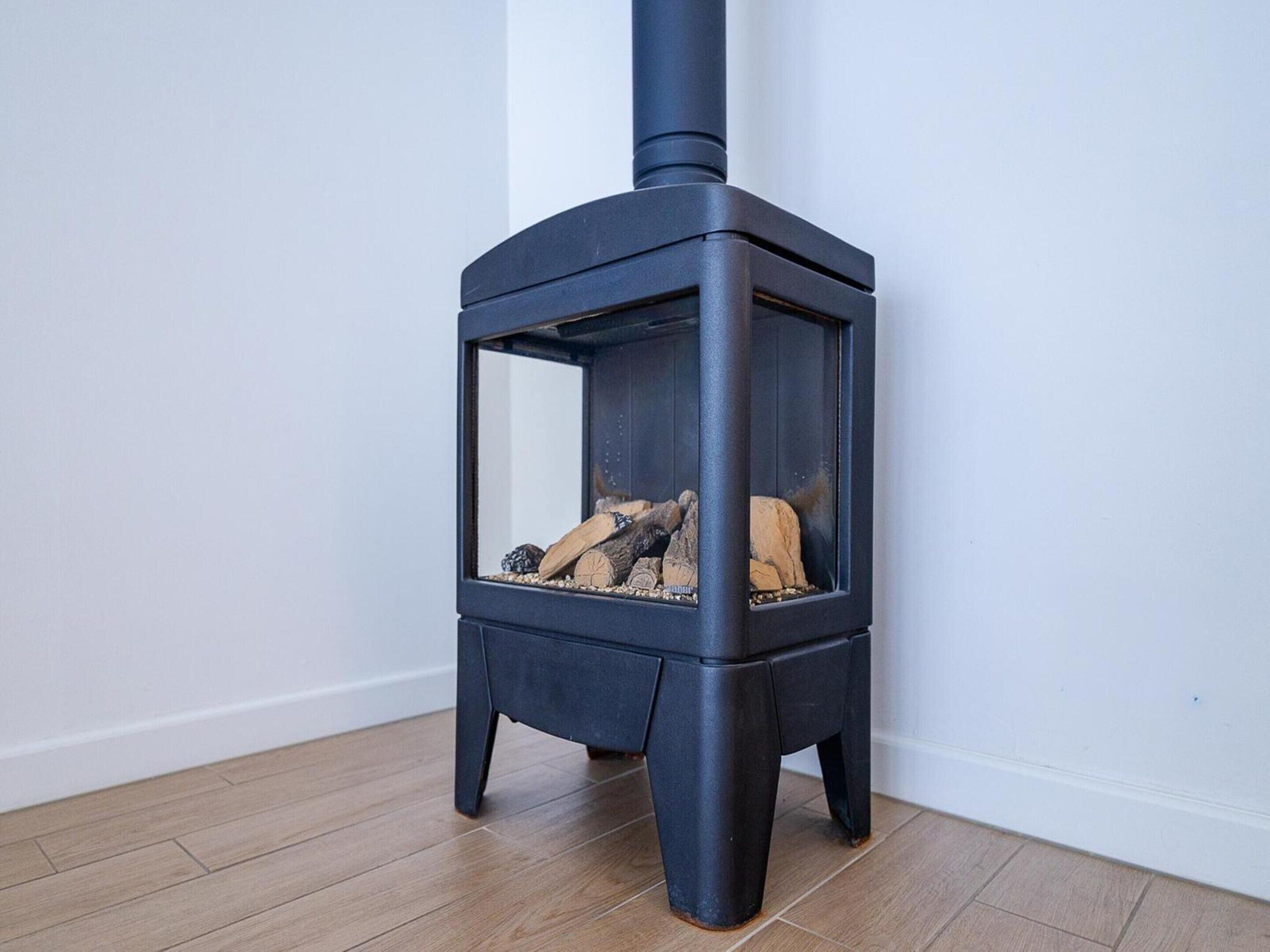 A black wood-burning stove with a glass door, placed on a wooden floor against a white wall.