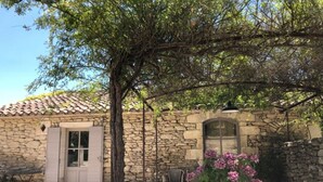 Un edificio de piedra con techo de tejas, una puerta y una ventana. Hay una planta en maceta con flores rosas frente al edificio.