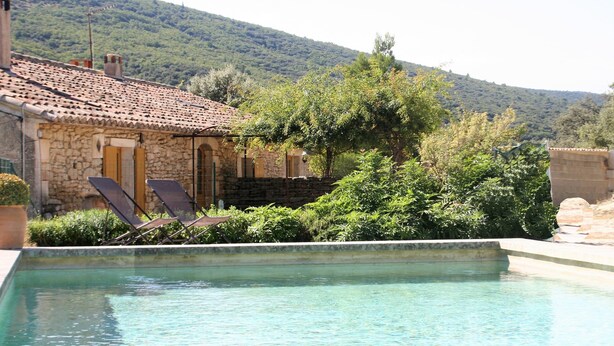 Un edificio de piedra con techo de tejas, piscina y un cerro al fondo.