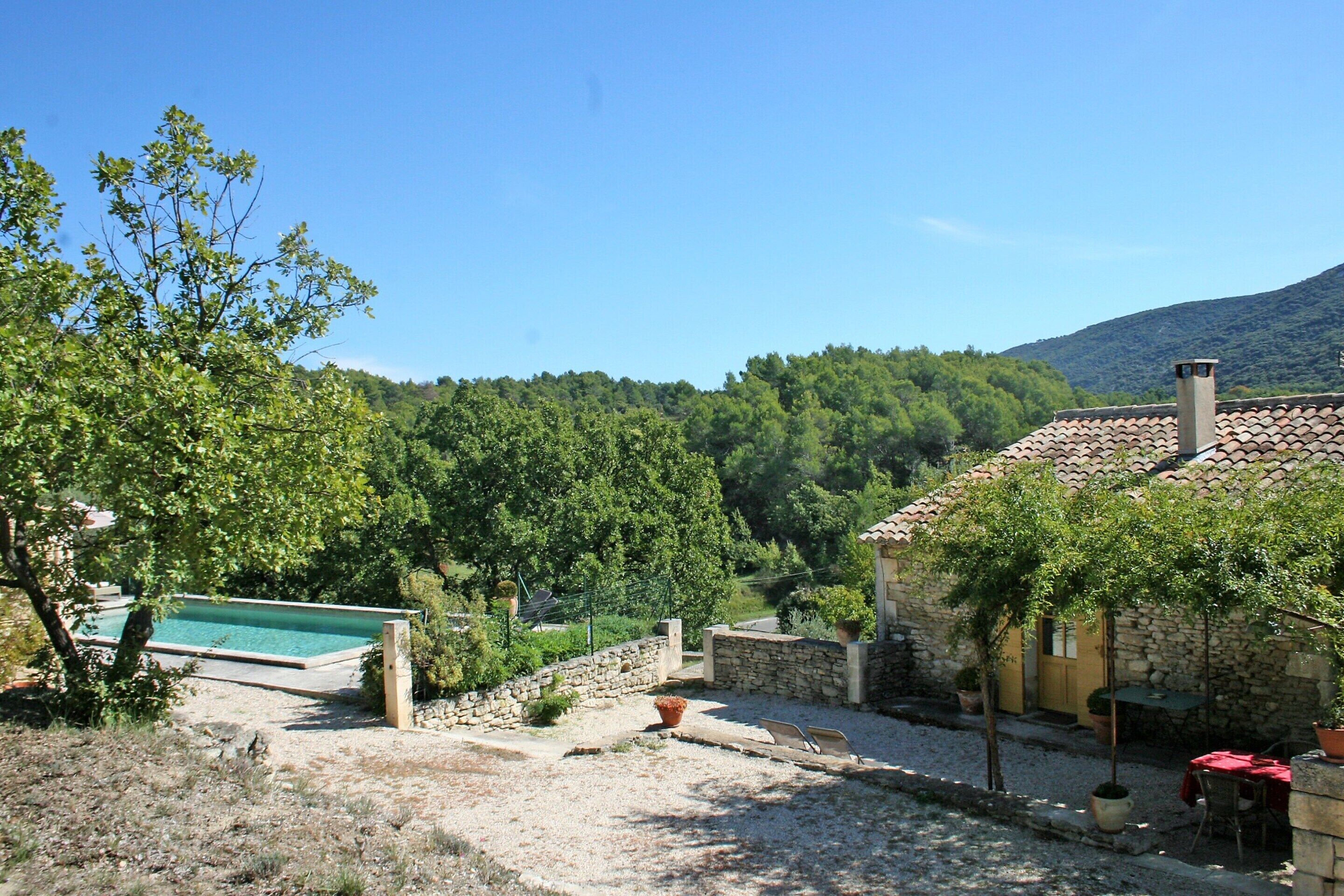 A stone house with a swimming pool, a patio with outdoor furniture, and a view of a forested hillside.