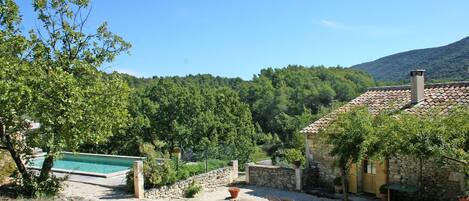 A stone house with a swimming pool, a patio with outdoor furniture, and a view of a forested hillside.