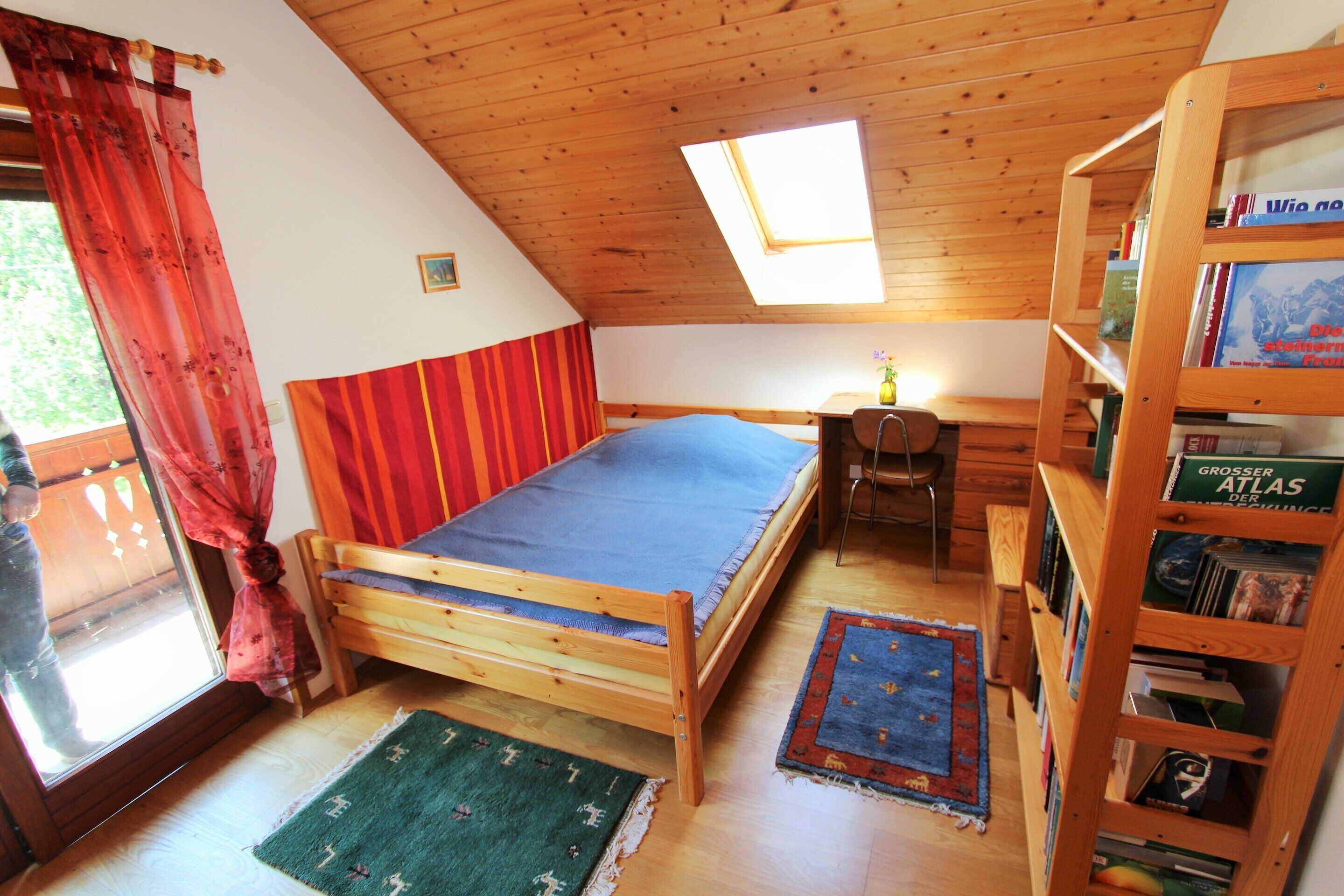 Ein Zimmer mit Holzboden, einem Bett mit blauer Matratze, einem Schreibtisch mit Stuhl und einem Bücherregal.