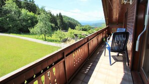 Un balcón con barandilla de madera, una silla y vistas a un paisaje verde con árboles y un camino.