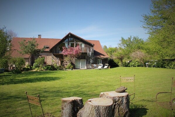 Une maison avec un toit recouvert de tuiles rouges, une grande pelouse devant et une terrasse en bois avec des chaises.