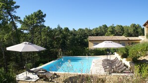 Una piscina con sillones de descanso y sombrillas, rodeada de árboles y un edificio con tejado de baldosas.