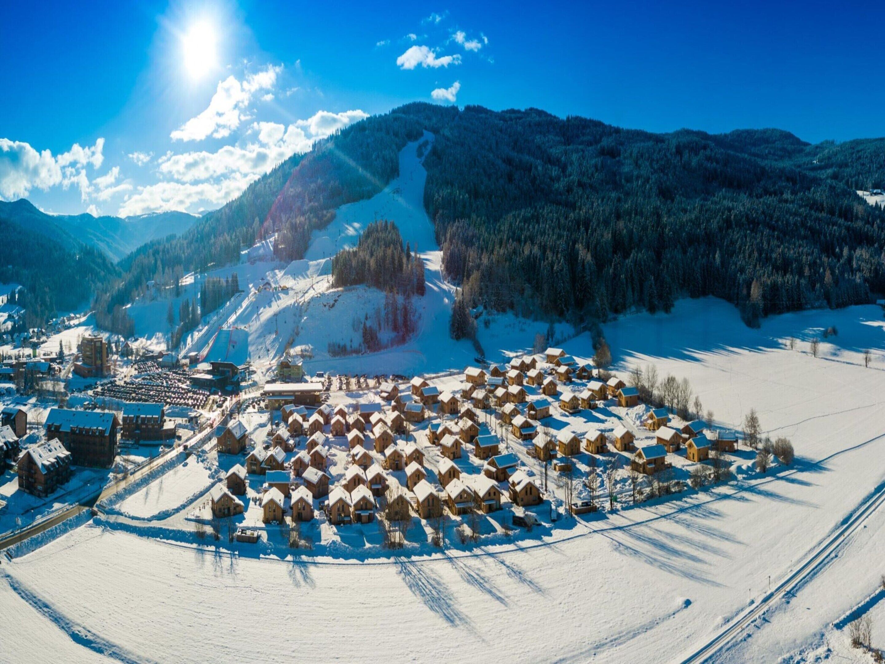 Ein verschneites Dorf mit Holzhäusern und einer Skipiste im Hintergrund.