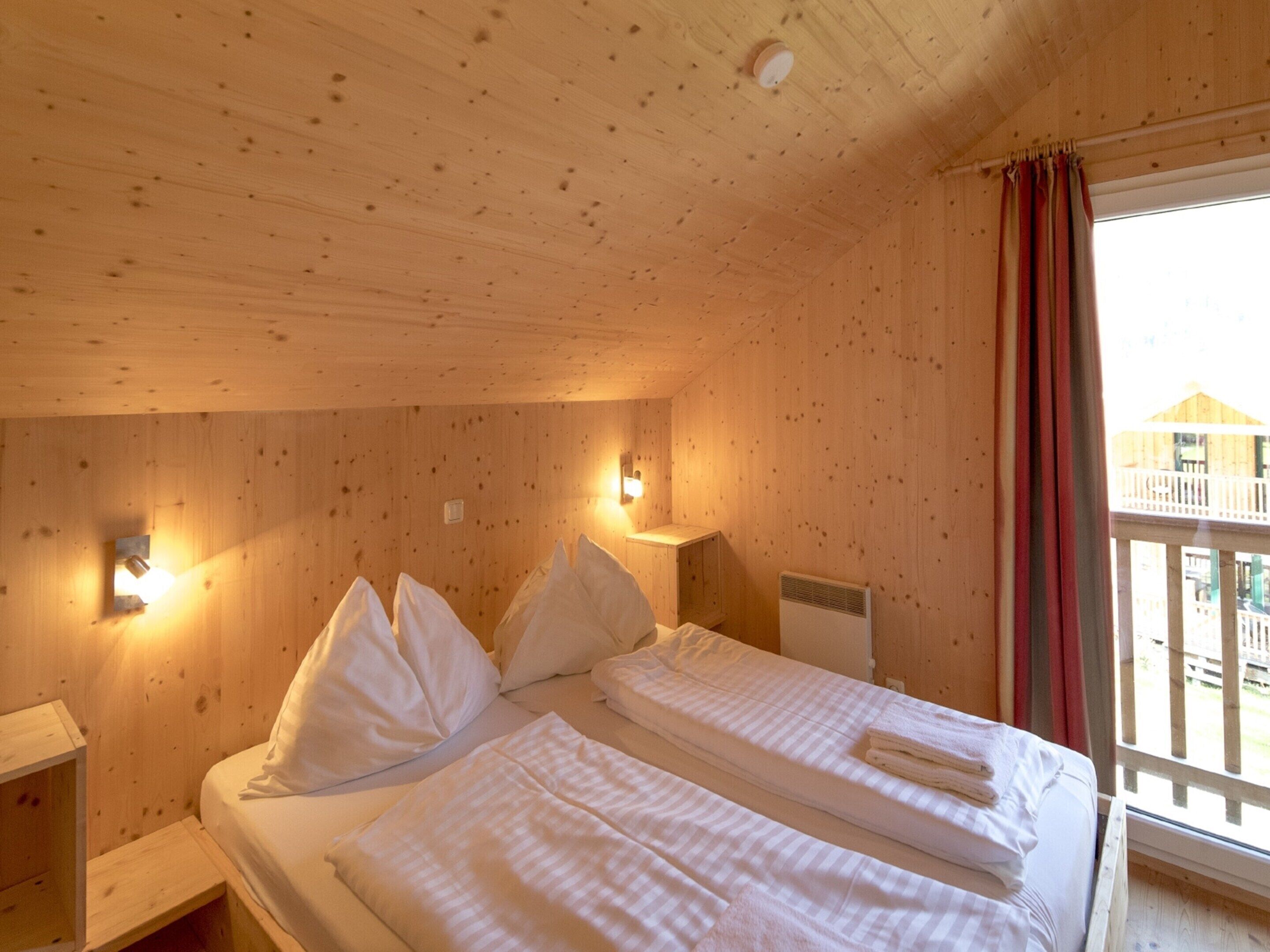 A wooden cabin room with a bed, two wall-mounted lights, a heater, and a window with a view of a balcony.