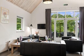 A modern living room with a sofa, a coffee table, and a flat-screen TV. There is a window with curtains, a radiator, and a small table with a lamp. - Kempen Comfort With Sauna & Tub (Vessem)