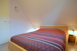 A bedroom with a sloped ceiling, a bed with a patterned bedspread, a bedside table with a lamp, and a wall-mounted light fixture. - Holiday Home in Heeten Near Forest (Heeten)