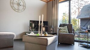 A modern living room with a sofa, ottoman, and a side table with candles. A large window provides a view of the outdoors. - Tranquil Holiday Home in Alphen-chaam With Stables (Alphen)