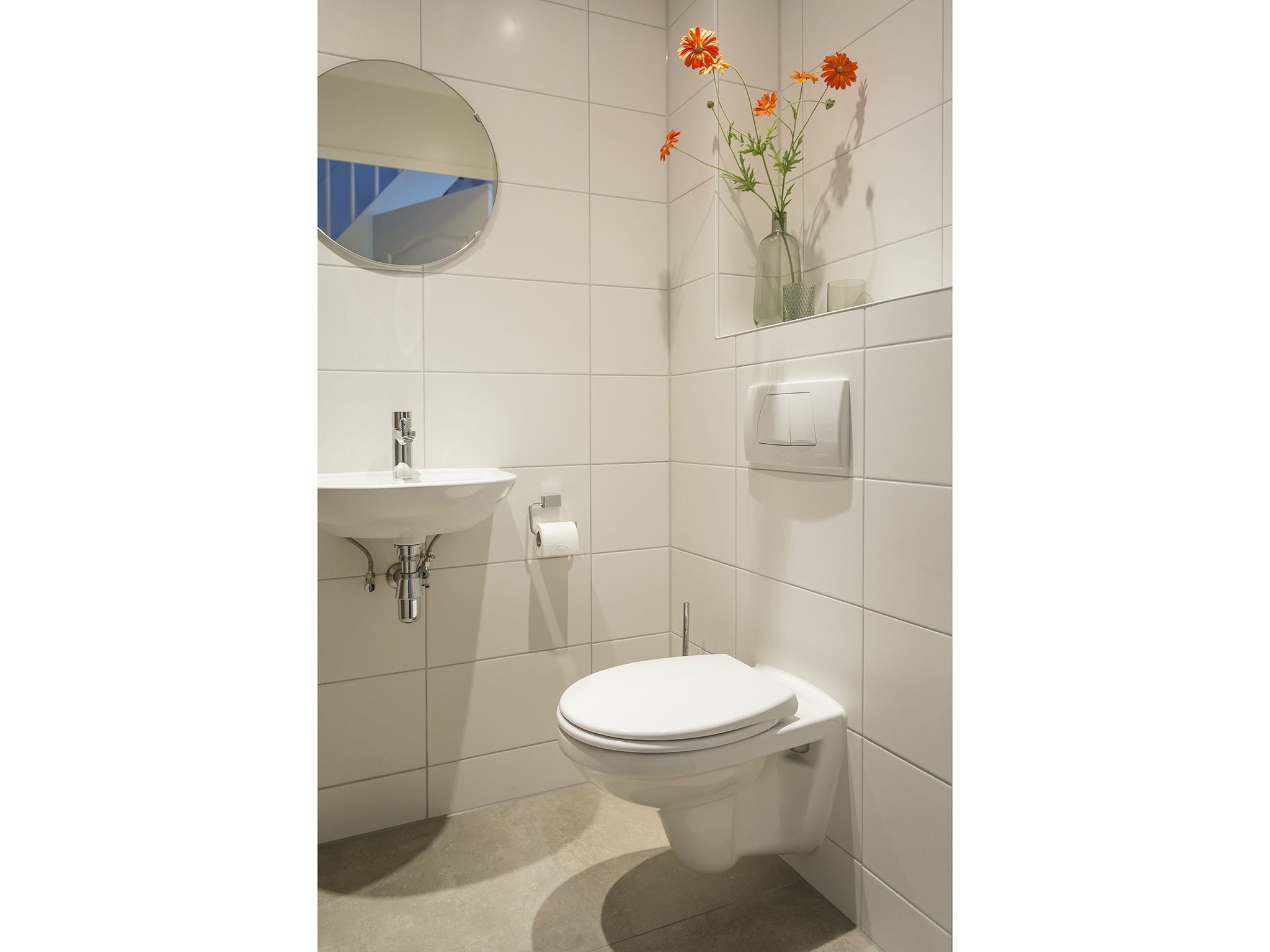 A modern bathroom with a white sink, toilet, and a mirror. There is a vase with flowers on the wall.