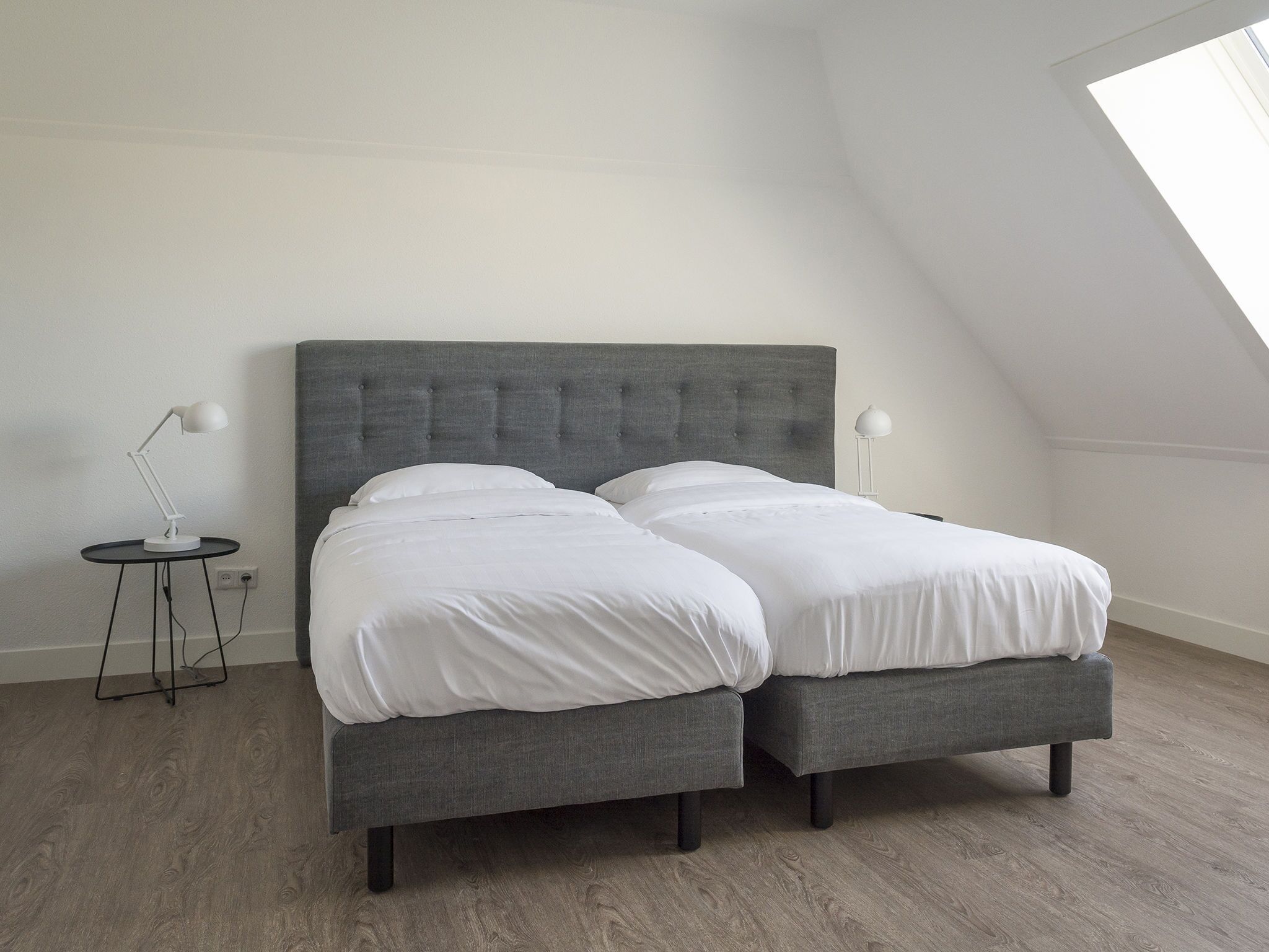 A modern bedroom with a grey upholstered bed, two bedside tables with lamps, and a sloped ceiling with a skylight.