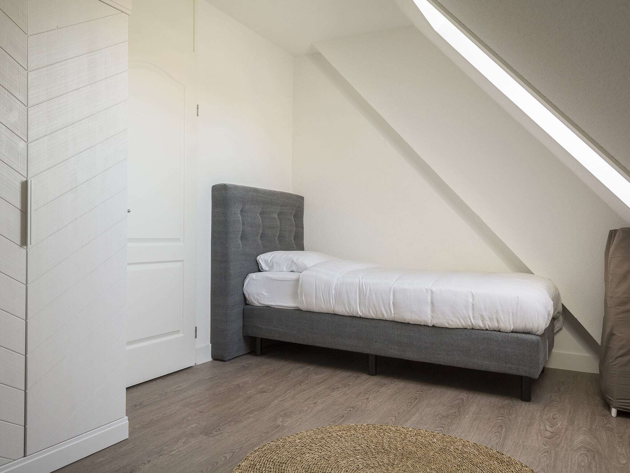 A bedroom with a sloped ceiling, a grey upholstered bed, a white door, and a wooden floor.