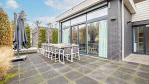 A modern outdoor patio with a dining table and chairs, large sliding glass doors, and a covered seating area. - Modern Villa in Harderwijk with Hot Tub (Zeewolde)