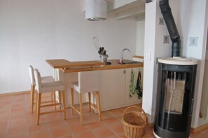 Een compacte keuken met een houten aanrecht, een spoelbak, twee witte stoelen, een rieten mand en een houtkachel.