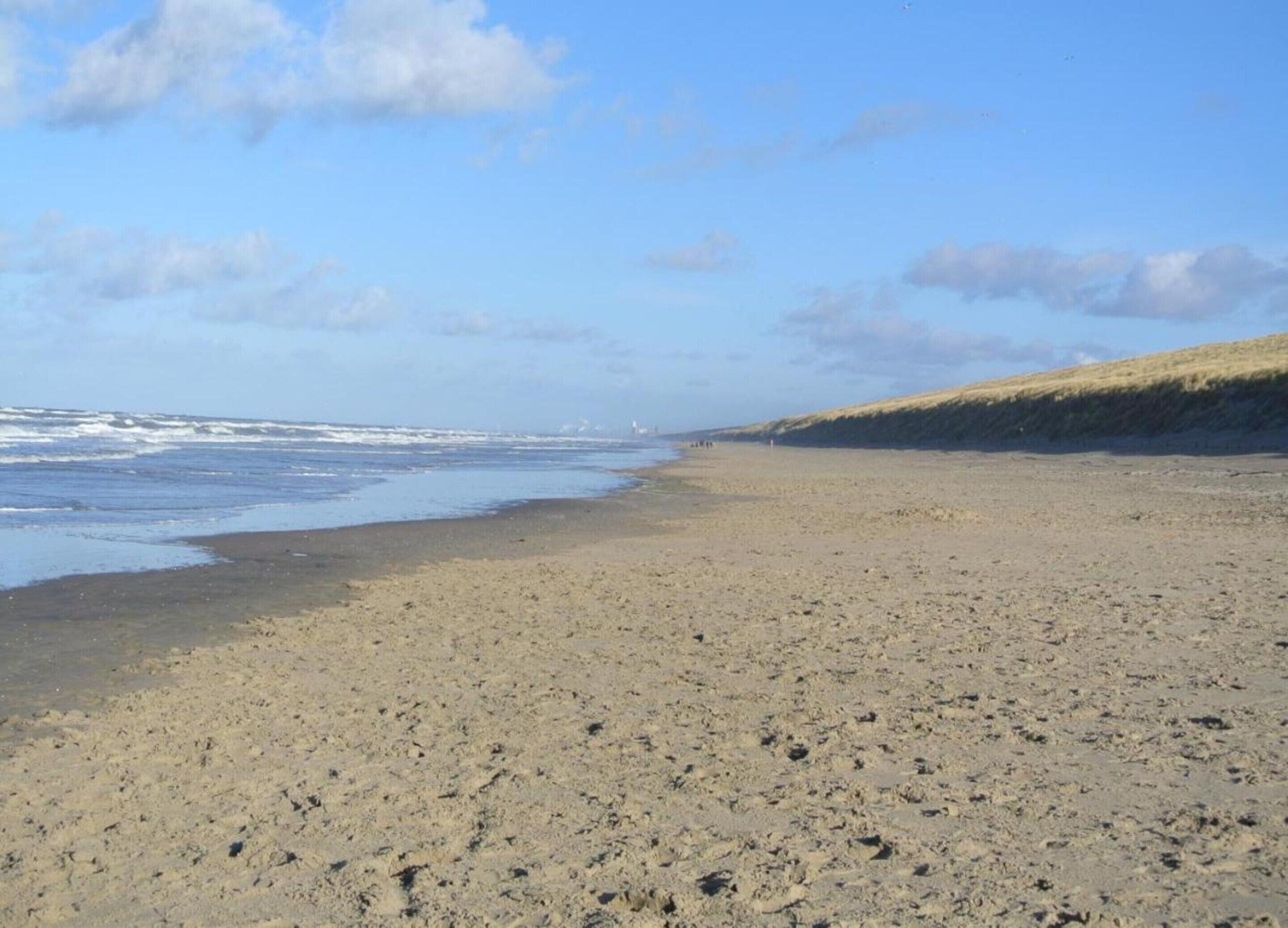 Uma extensa praia de areia com ondas suaves, céu claro e uma colina gramada ao fundo.