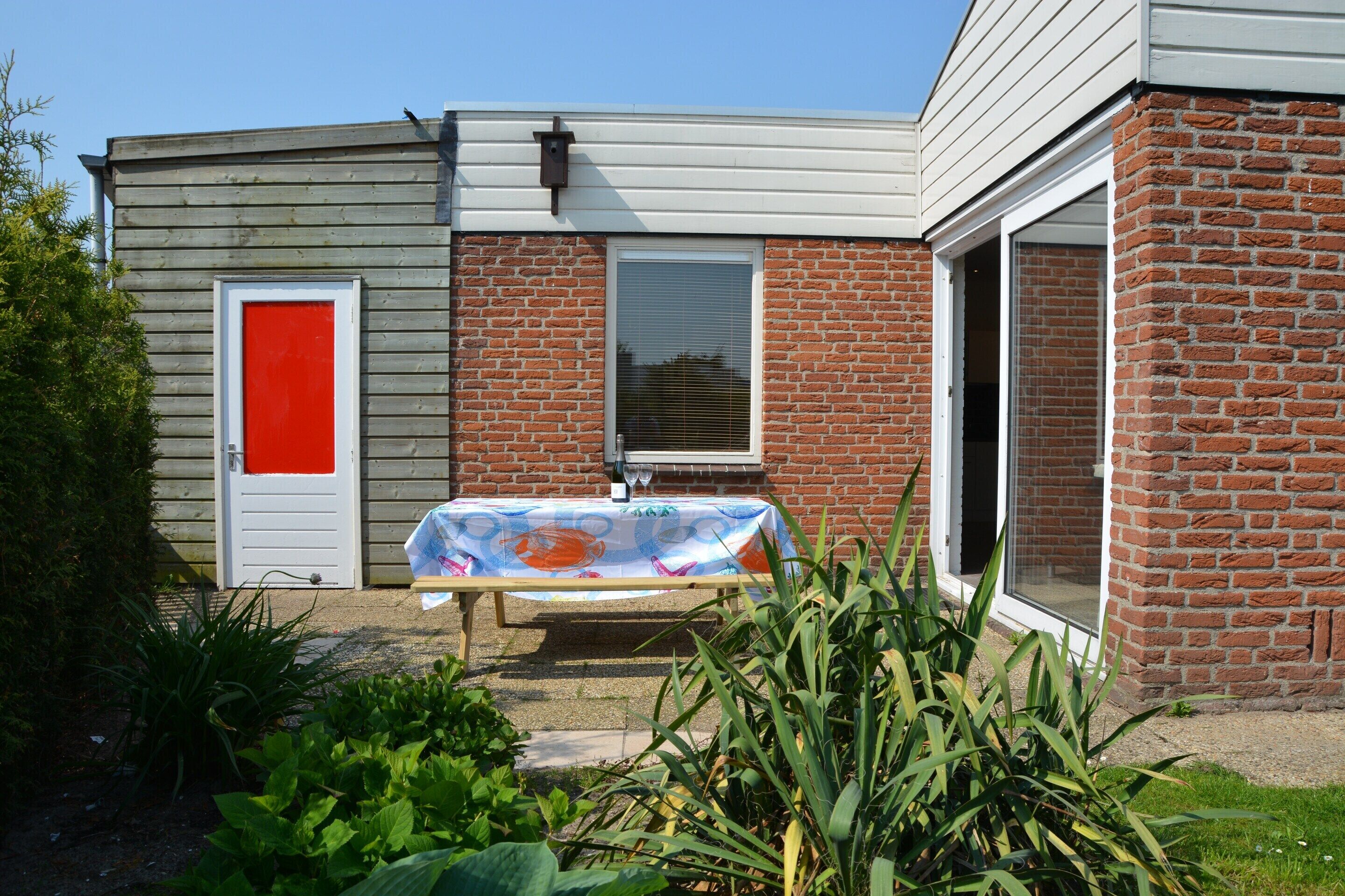 Ein Haus mit roter Tür, einer überdachten Terrasse mit Tisch und einer Ziegelaußenwand.