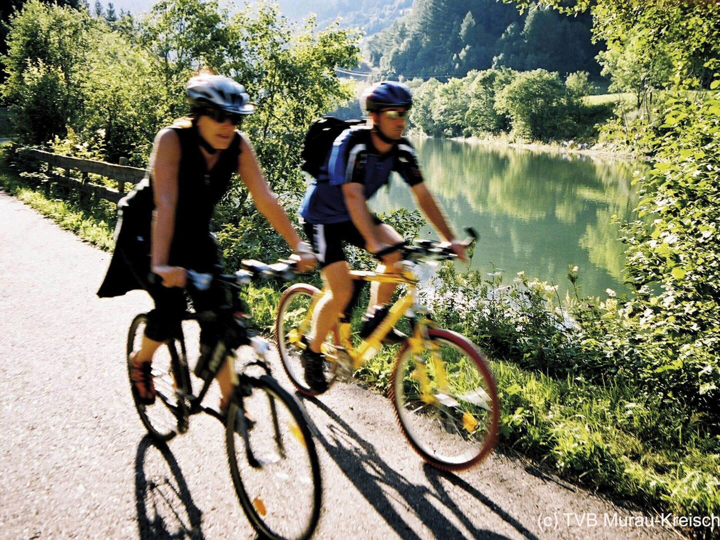 Zwei Radfahrer auf einem asphaltierten Weg, im Hintergrund ein See und viel Grün.
