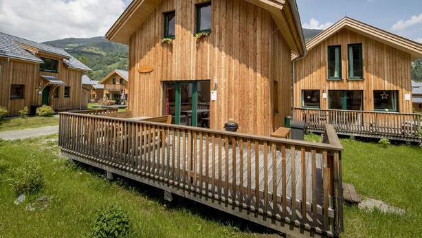 Una baita di legno con una terrazza, immersa nel verde e con montagne sullo sfondo.